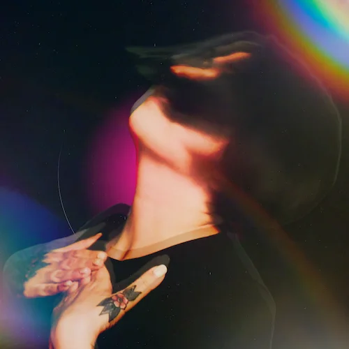 a photo of a woman with her hands on her heart, looking up and to the left. She is blurry and artifacted, against a space-like backdrop containing a pink orb. At the top right and bottom left there are rainbow-like lens flares, making it seem like she is in space.